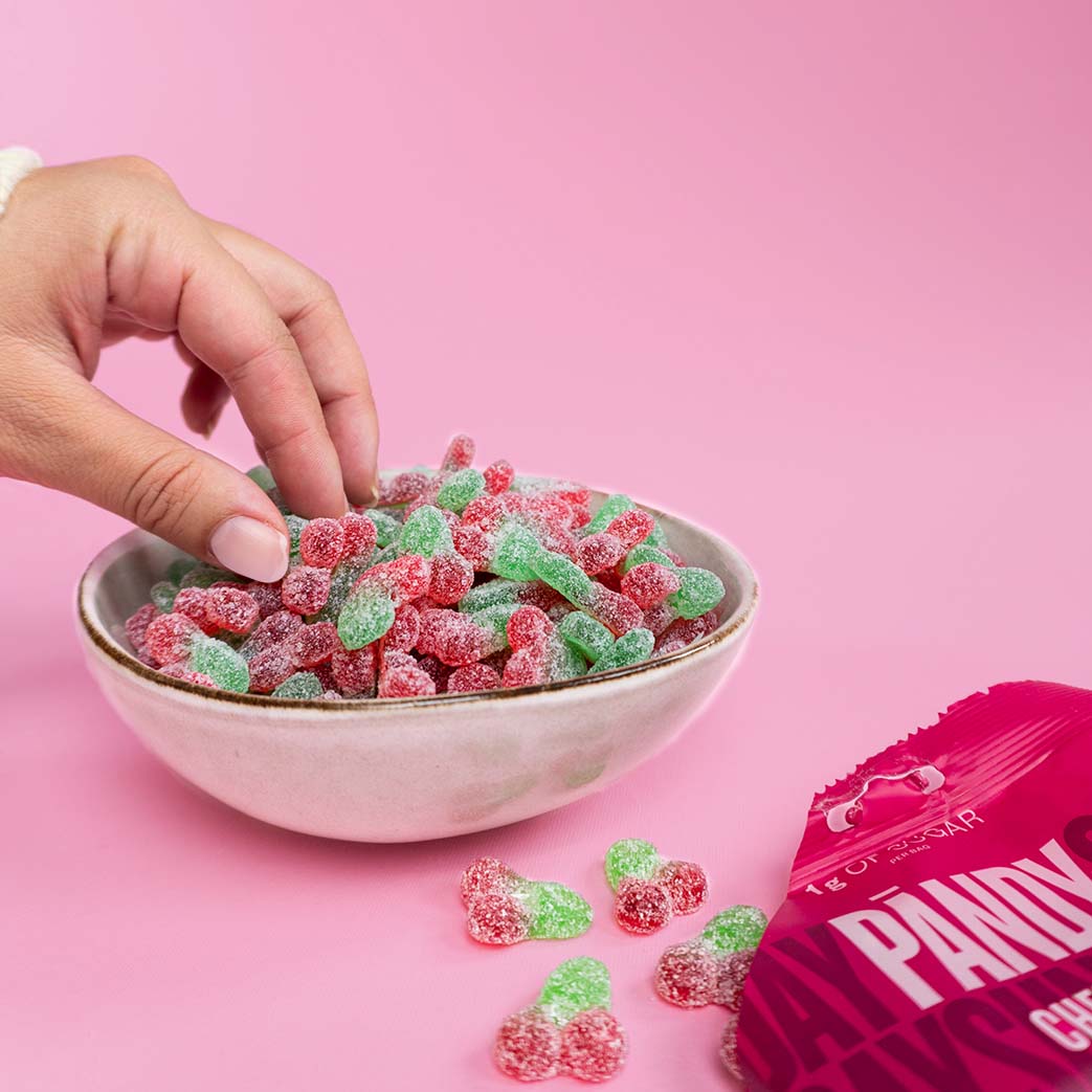 A hand reaches into a small bowl of sugar-coated cherry-shaped candies in pink and green, with a pink bag labeled Pandy Candy visible on the right against a solid pink background.